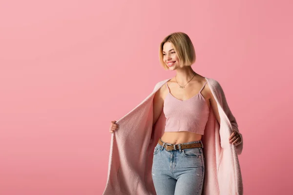 Mujer joven y complacida en cárdigan mirando hacia otro lado aislado en rosa - foto de stock