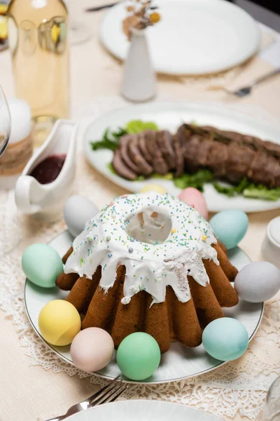 Вкусный пасхальный торт с окрашенными яйцами рядом с соусом и жареным мясом на размытом фоне — стоковое фото