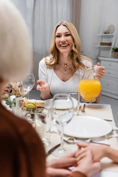 Cheerful woman holding jug of orange juice while talking to blurred mother during easter dinner — Stock Photo