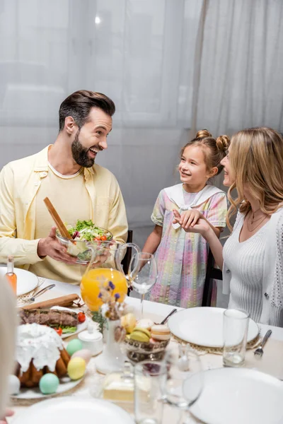 Aufgeregter Mann hält Schüssel mit Gemüsesalat in der Nähe von Tochter und Frau beim Osteressen — Stockfoto