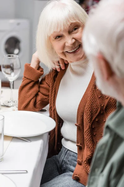 Старша жінка посміхається за розмитим чоловіком, сидячи за святковим столом — стокове фото