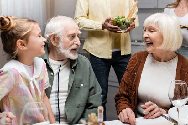 Senior woman laughing near husband and granddaughter during family dinner — Stock Photo