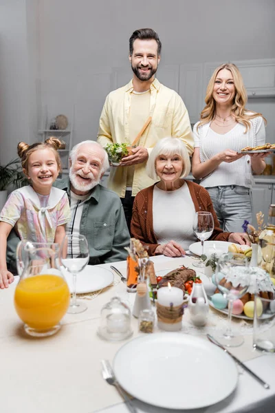 Familia feliz sonriendo a la cámara cerca de la mesa servida con cena de Pascua - foto de stock