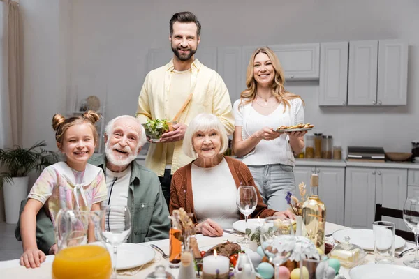 Coppia felice che tiene l'insalata vegetale e biscotti vicino a famiglia durante celebrazione pasquale — Foto stock