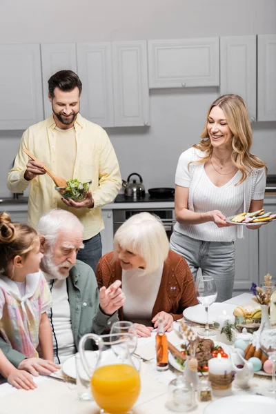 Uomo sorridente mescolando insalata di verdure durante la cena di Pasqua in famiglia — Foto stock