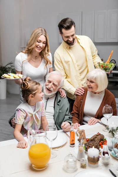 Happy couple holding vegetable salad and cookies near girl and happy grandparents — Stock Photo