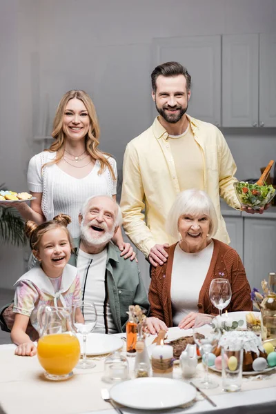 Casal sorridente com salada de legumes e biscoitos olhando para a câmera perto da família em casa — Fotografia de Stock