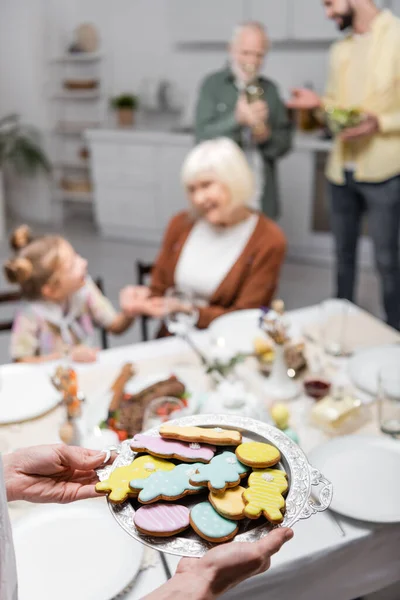 Женщина держит поднос с традиционными пасхальными печеньями рядом размытой семьи на кухне — стоковое фото