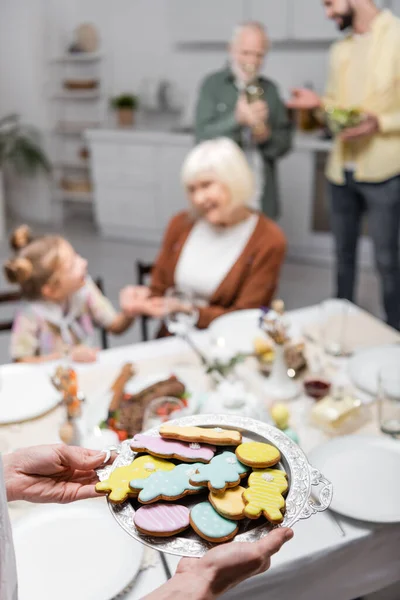 Woman holding tray with traditional easter cookies near blurred family in kitchen — Stock Photo