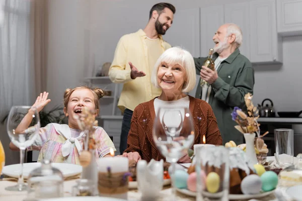 Excitada chica agitando la mano cerca de la abuela sonriente durante la cena festiva en casa - foto de stock