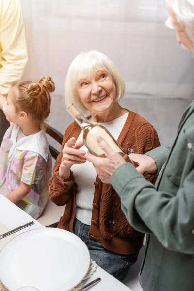 Sorrindo mulher idosa apontando para garrafa de vinho durante o jantar de Páscoa com a família — Fotografia de Stock