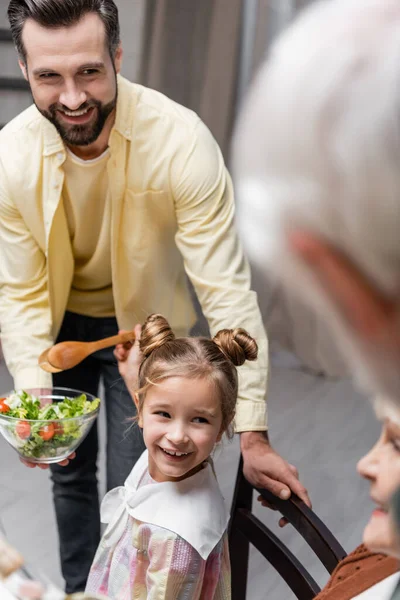 Homem sorrindo segurando salada de legumes perto de filha alegre e família turva — Fotografia de Stock