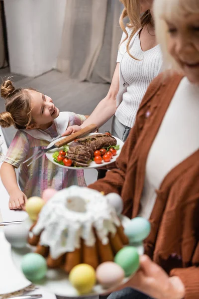 Fille heureuse souriant près de la mère et la mamie floue tenant gâteau de Pâques et de la viande avec des légumes — Photo de stock