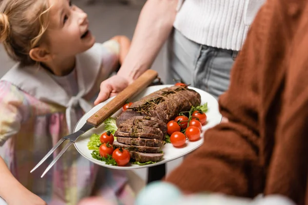 Mujer sosteniendo plato con carne y verduras frescas cerca de hija excitada - foto de stock