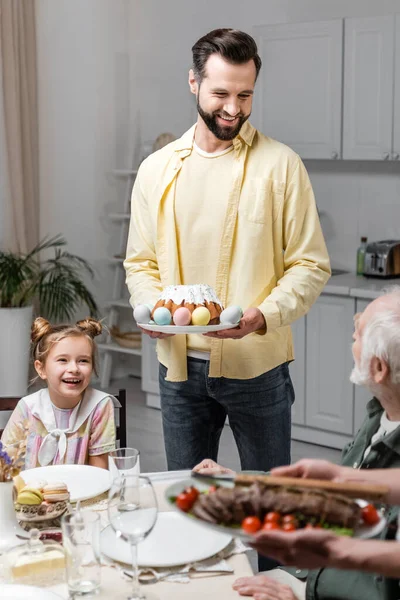 Улыбающийся мужчина с пасхальным тортом и раскрашенными яйцами рядом с семьей во время праздничного ужина — стоковое фото