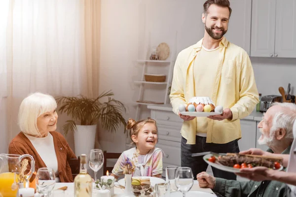Счастливый человек с пасхальным тортом и окрашенные яйца во время пасхального ужина с семьей — стоковое фото