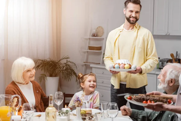 Hombre feliz con pastel de Pascua y huevos pintados durante la cena de Pascua con la familia - foto de stock