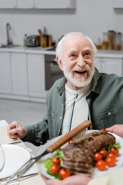 Uomo anziano stupito guardando moglie che tiene piatto con carne durante la celebrazione pasquale — Foto stock