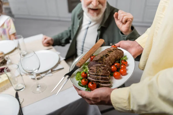 Vista recortada del hombre sosteniendo carne con verduras frescas cerca del padre mayor durante la cena de Pascua - foto de stock