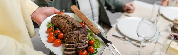 Vue recadrée de l'homme tenant assiette avec de la viande et des légumes frais près de la famille floue, bannière — Photo de stock