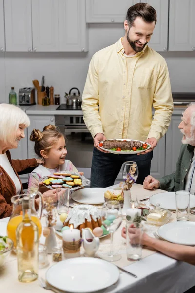 Felice uomo che tiene piatto con carne vicino alla famiglia che celebra Pasqua — Foto stock