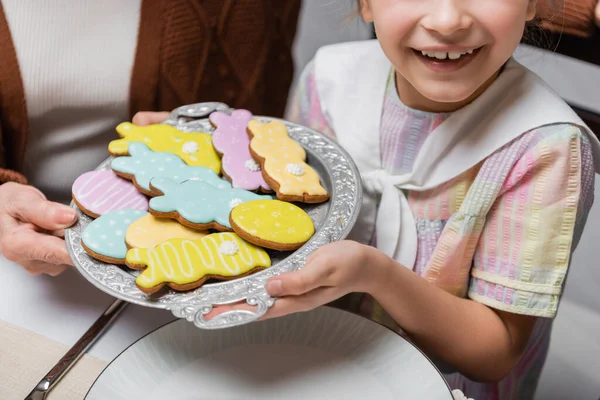 Vista recortada de niña sonriente sosteniendo bandeja con coloridas galletas de Pascua - foto de stock