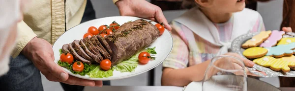 Vista parcial del hombre sosteniendo plateado con carne, tomates cherry y lechuga cerca de hija con galletas, pancarta - foto de stock