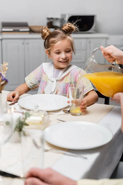Niña preadolescente sonriendo cerca de la madre vertiendo jugo de naranja durante la cena de Pascua — Stock Photo