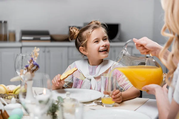 Fröhliches Mädchen mit traditionellem Osterkeks in der Nähe von Mama, die frischen Orangensaft einschenkt — Stockfoto