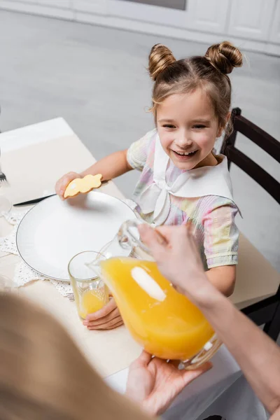 Femme floue versant du jus d'orange près de fille heureuse tenant biscuit de Pâques — Photo de stock