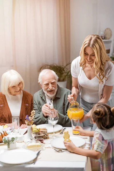 Sonriente mujer vertiendo jugo de naranja mientras familia teniendo cena de Pascua en casa - foto de stock