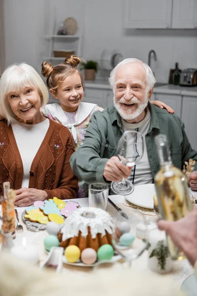 Happy man holding glasses near bottle of wine during easter dinner with family — Stock Photo