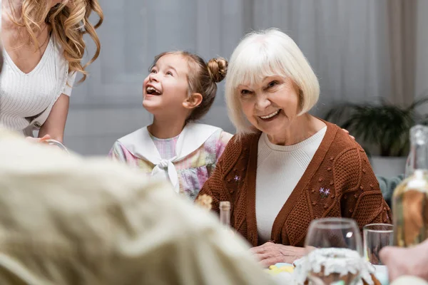 Mujer mayor sonriendo cerca nieta sorprendida durante la cena festiva con la familia - foto de stock