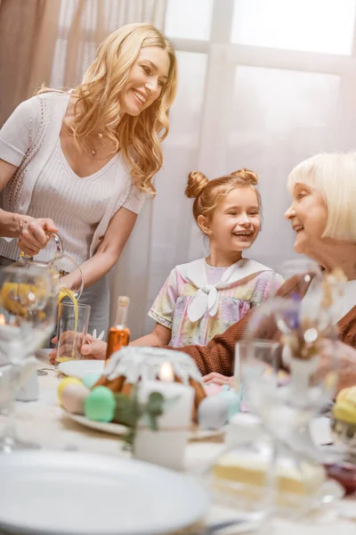 Cheerful woman pouring orange juice near senior mom and happy daughter — Stock Photo