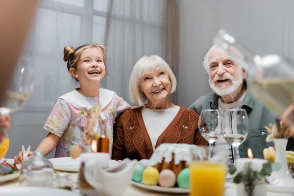 Cheerful girl smiling near happy grandparents and festive dinner on table — Stock Photo