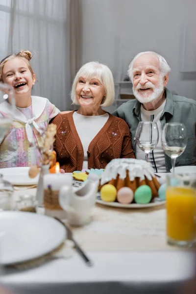 Chica alegre con abuelos cerca de pastel de Pascua y huevos pintados en la mesa servida - foto de stock