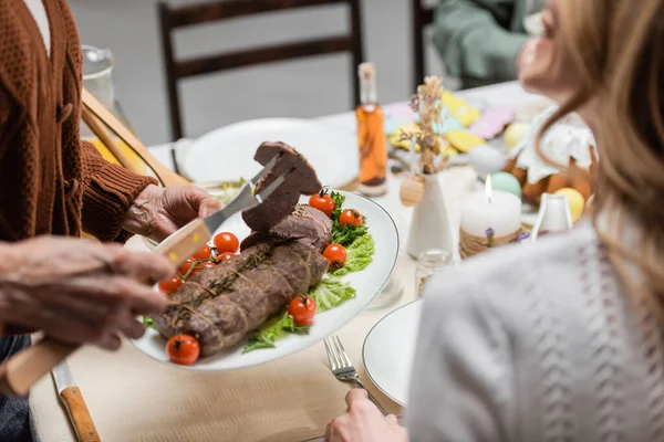 Vista recortada de la mujer mayor sirviendo carne cerca de hija adulta durante la cena de Pascua - foto de stock
