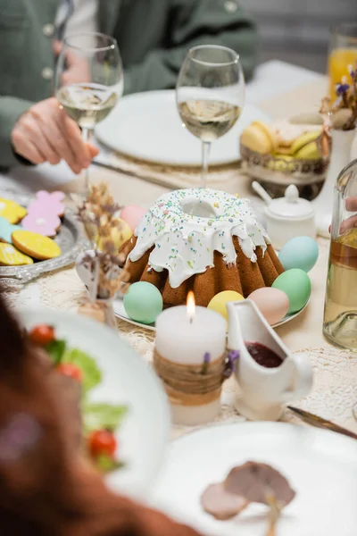 Vista parcial del hombre cerca de delicioso pastel de Pascua, huevos pintados y copas de vino en la mesa festiva - foto de stock