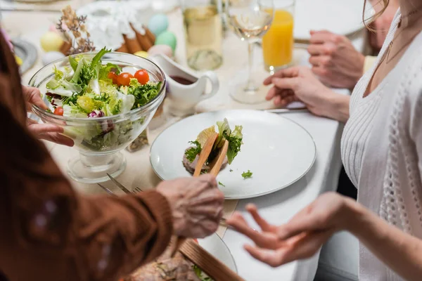 Обрізаний вид жінки, що тримає миску зі свіжим овочевим салатом біля розмитої сім'ї, що вечеряє на Великдень — стокове фото