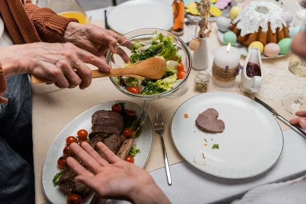 Vista parcial de la mujer mayor sosteniendo ensalada de verduras cerca de la carne en la mesa festiva — Stock Photo