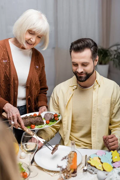 Seniorin serviert gebratenes Fleisch beim Osteressen mit erwachsenem Sohn — Stockfoto