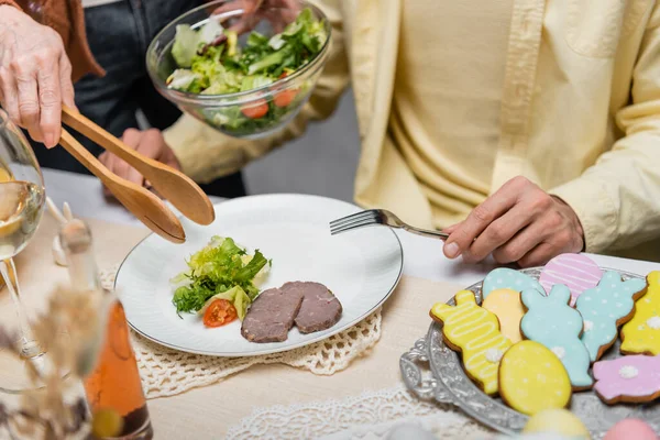 Vista parcial de la mujer mayor sosteniendo fórceps de servicio y ensalada de verduras cerca de hijo adulto - foto de stock