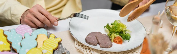 Vue recadrée de l'homme à la fourchette et de la femme à la pince de service près de biscuits colorés, salade et viande, bannière — Photo de stock