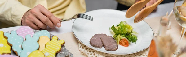 Vista recortada del hombre con tenedor y la mujer con pinzas de servir cerca de galletas de colores, ensalada y carne, pancarta — Stock Photo