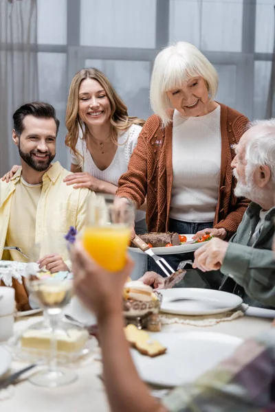 Donna anziana sorridente che serve carne fritta vicino alla famiglia e bambino sfocato con vetro di succo d'arancia — Foto stock