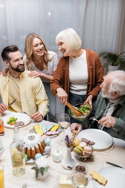 Пожилая женщина держит миску с овощным салатом рядом с счастливой семьей — стоковое фото