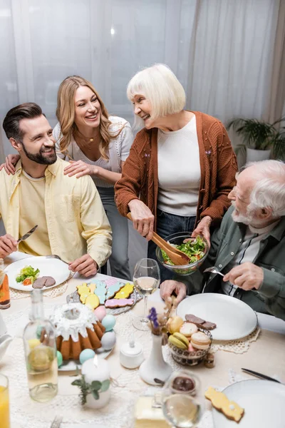 Mulher sênior segurando tigela com salada de legumes perto da família feliz — Fotografia de Stock