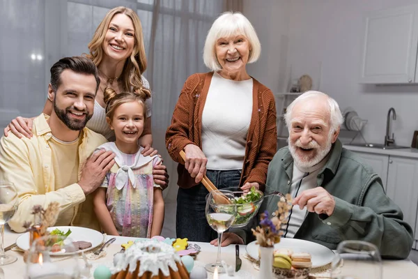 Familia alegre mirando la cámara cerca de la mesa servida con cena festiva de Pascua - foto de stock