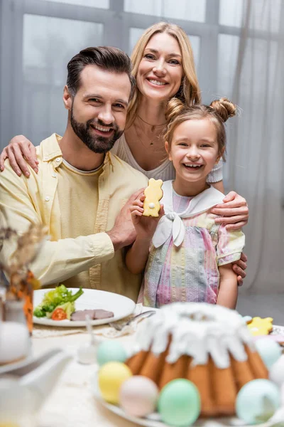 Cheerful girl holding traditional easter cookie during dinner with happy parents — Stock Photo