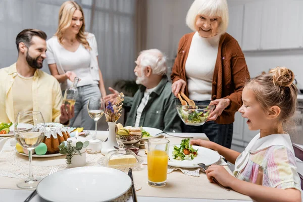 Mulher sênior segurando salada de legumes perto de neta e família turva — Fotografia de Stock